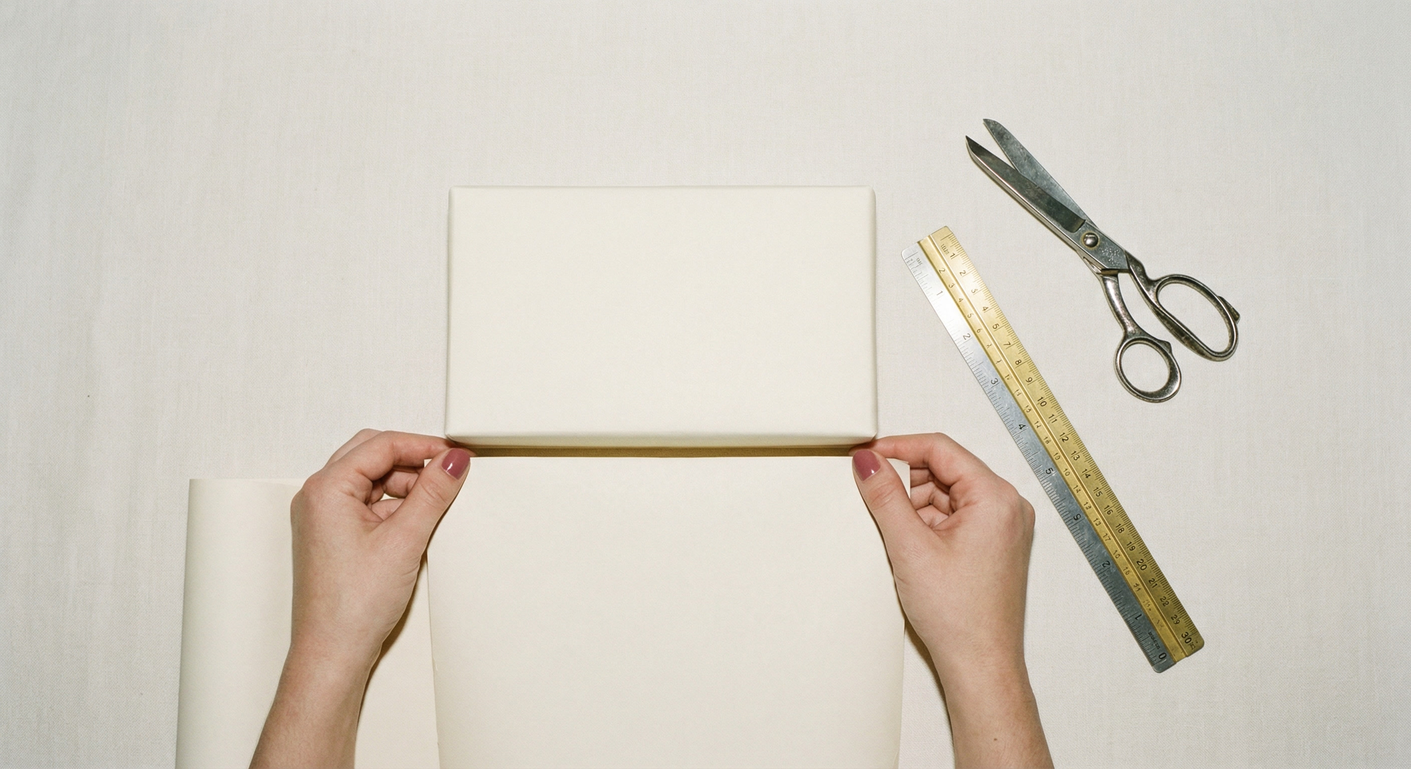 Top-down view of hands measuring cream wrapping paper against a rectangular gift box with scissors and a ruler nearby