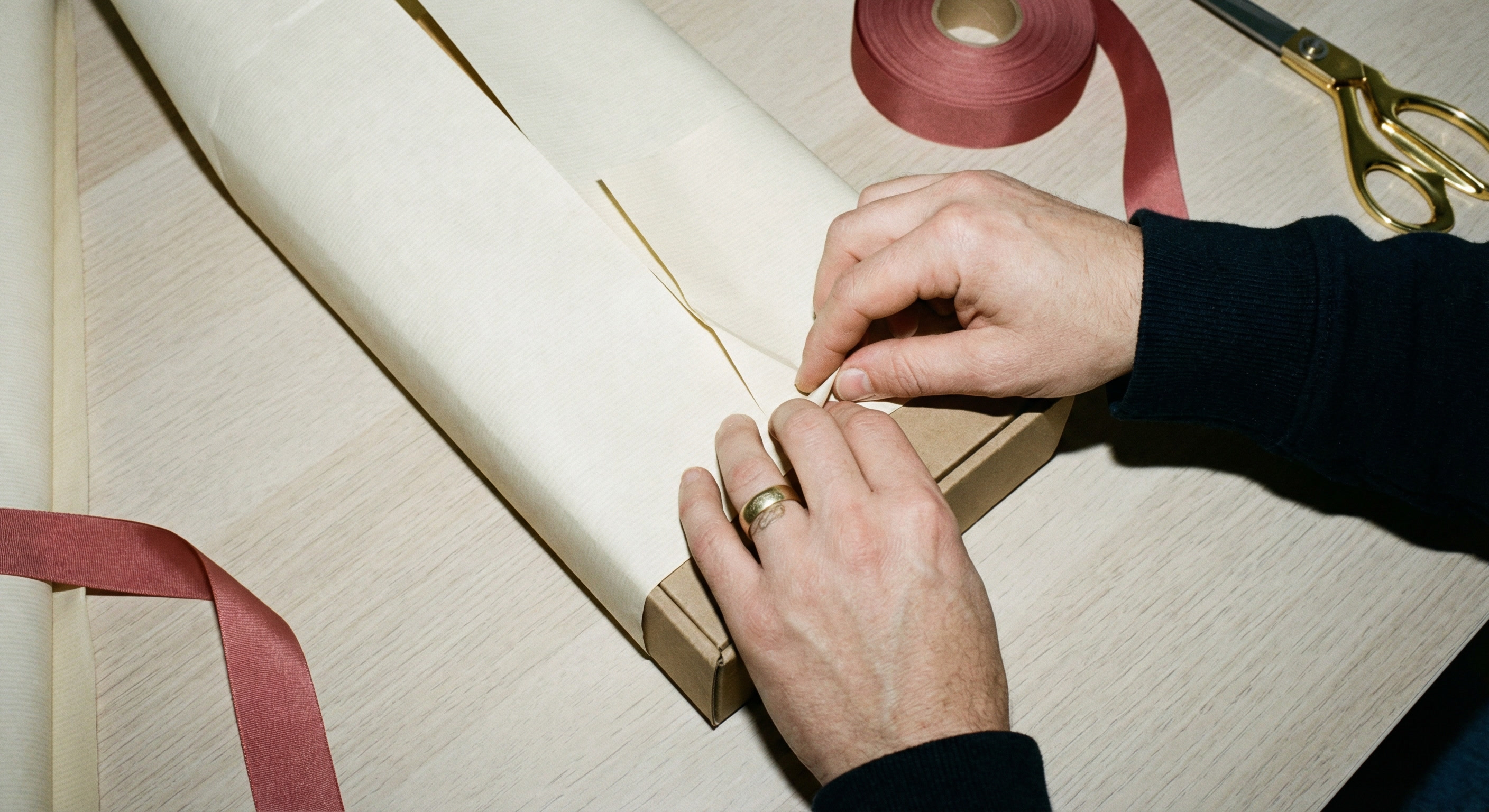 Close-up top-down view of hands folding the first long edge of cream wrapping paper neatly over a rectangular box