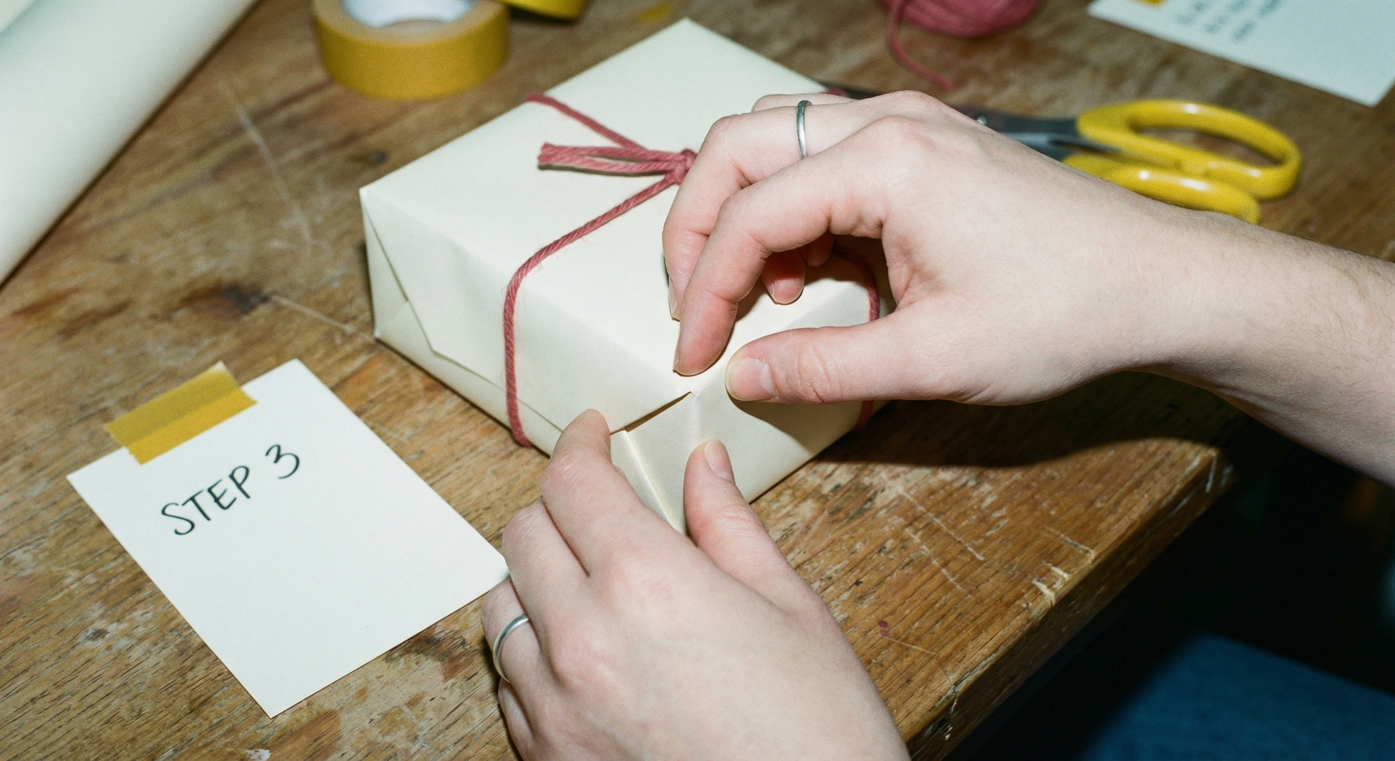 Top-down close-up of hands pinching and folding wrapping paper at the corner of a box to create a sharp triangular flap